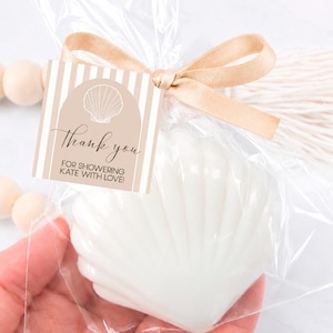 May include: A white shell-shaped item packaged in a clear plastic bag with a beige ribbon. A square tag is attached, featuring a beige and white striped pattern, an arch, a shell illustration, and the text: Thank you FOR SHOWERING KATE WITH LOVE!