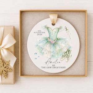 May include: A round white ceramic ornament featuring a watercolor illustration of a light green and blue tutu dress, ballet slippers, and a wand. Text reads THE NUTCRACKER 2026, Amelia, and AS THE DEW DROP FAIRY. A gold ribbon loop is attached at the top.