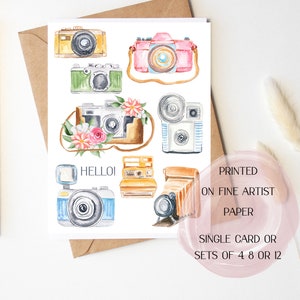 May include: A set of eight watercolor illustrations of vintage cameras, including a yellow camera, a pink camera, a green camera, a brown camera with flowers, a white camera, a blue camera with a flash, a yellow Polaroid camera, and a brown camera with a bellows. The text "HELLO!" is printed on the card. The card is printed on fine artist paper and is available as a single card or in sets of 4, 8, or 12.