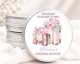 May include: A round silver metal tin with a white label featuring a watercolor illustration of two vintage suitcases adorned with pink flowers. The text reads TRAVELING FROM MISS TO MRS! and Melissa's WEDDING SHOWER in pink and black lettering.