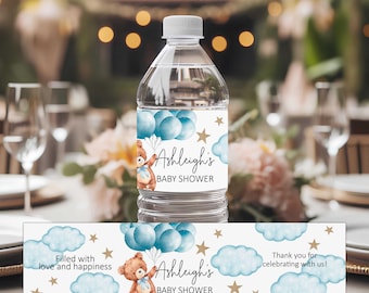 May include: A clear plastic water bottle with a white label featuring a brown teddy bear holding blue balloons, gold stars, and blue clouds. The text reads Ashleigh's BABY SHOWER, Filled with love and happiness, and Thank you for celebrating with us.