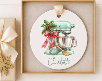 May include: A round white ceramic ornament featuring a light green stand mixer illustration decorated with a red bow, holly, a whisk, and a wooden spoon. The name Charlotte is printed in green script below the mixer. The ornament hangs by a gold ribbon.