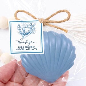 May include: A blue seashell-shaped soap packaged in a clear plastic bag tied with a brown twine bow. A square white tag with a blue border features a blue illustration of a seashell and the text Thank you FOR SHOWERING MADISON WITH LOVE!