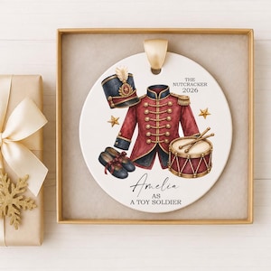 May include: A round white ceramic ornament featuring a red and navy toy soldier uniform, a drum, and a hat. Text reads: THE NUTCRACKER 2026, Amelia AS A TOY SOLDIER. The ornament has a gold ribbon loop and is presented in a gold-colored gift box.