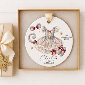 May include: A round white ceramic ornament featuring a gray and cream tutu dress, mouse ears with a red bow, ballet slippers, a gift box, and gold stars. Text reads: THE NUTCRACKER 2026 and Charlotte AS A MOUSE. The ornament has a gold ribbon hanger.