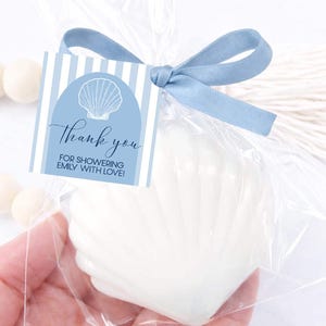 May include: A white seashell-shaped item packaged in a clear plastic bag tied with a dusty blue ribbon. A square tag features a blue and white striped pattern, a white seashell illustration, and the text Thank you FOR SHOWERING EMILY WITH LOVE!