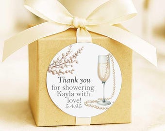 May include: A square tan gift box tied with a cream-colored satin ribbon. A circular sticker on the front features a champagne flute, a string of pearls, and the text: Thank you for showering Kayla with love! 5.4.25. The design includes pearl-like branches.