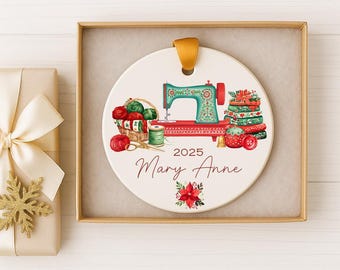 May include: A round white ceramic ornament featuring a teal sewing machine, a basket of yarn, and a stack of fabric. The text reads 2025 and Mary Anne, with a poinsettia graphic below. It hangs from a gold ribbon inside a square gift box.