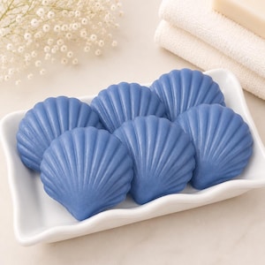 May include: A collection of six blue seashell-shaped soaps arranged in a white rectangular ceramic dish with wavy edges. Each soap features a detailed ribbed texture mimicking the natural appearance of a scallop shell, presented in a group.