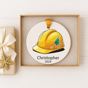 May include: A round white ceramic ornament featuring a yellow hard hat graphic with a small decorated Christmas tree on the side. The text "Christopher" and "2024" is printed in black below the hard hat. It has a gold ribbon loop for hanging.