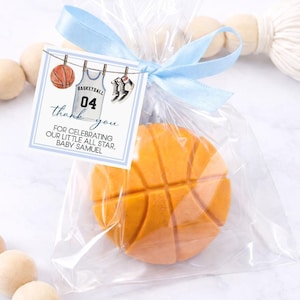 May include: A round orange basketball-shaped cookie inside a clear plastic bag tied with a light blue ribbon. A square tag attached to the bag features a basketball, a jersey with the number 04, and text reading: thank you FOR CELEBRATING OUR LITTLE ALL STAR, BABY SAMUEL.