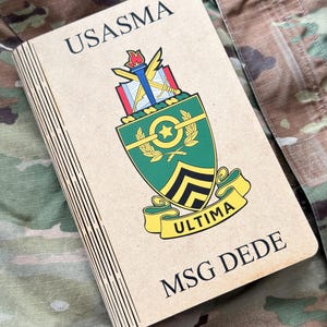 May include: A tan notebook with the text "USASMA" and "MSG DEDE" printed on the cover. The notebook features a colorful emblem with a torch, stars, and the word "ULTIMA" on a yellow banner. The background is camouflage fabric.