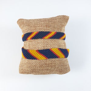 Puede incluir: Dos pulseras de amistad tejidas con rayas amarillas, rojas y azules sobre un fondo de arpillera marrón.