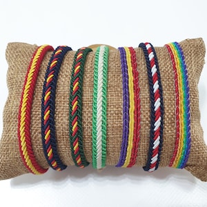 Può includere: Un set di otto braccialetti intrecciati colorati, esposti su un cuscino di juta. I braccialetti sono realizzati con cordoncino intrecciato in vari colori, tra cui rosso, giallo, verde, blu, bianco e nero.