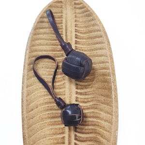 May include: Two brown leather keychains with knotted leather balls. The keychains are displayed on a light brown ceramic leaf.