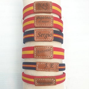 May include: Five red, yellow and blue striped bracelets with brown leather tags. The tags are engraved with the text "N&S", "Papa eres el mejor", "Sergio", "Antonio", and "J.E".