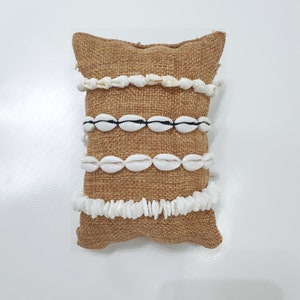 Può includere: Quattro braccialetti di conchiglie bianche su un cuscino di juta marrone. I braccialetti sono fatti di conchiglie di diverse dimensioni e forme.