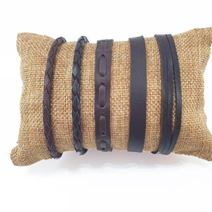 Può includere: Quattro braccialetti in pelle marrone su un cuscino di juta. I braccialetti sono di diversi stili, tra cui intrecciati, in rilievo e semplici.