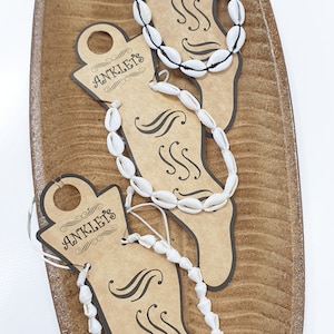 Könnte beinhalten: Drei weiße Muschelanhänger mit schwarzen Schnüren auf braunen Papieraushängeschildern mit der Aufschrift "Anklets". Die Schilder befinden sich auf einer braunen Holzoberfläche.