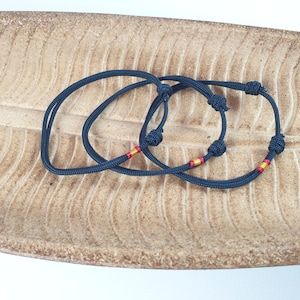 May include: Two black cord bracelets with red and gold accents. The bracelets are knotted and are lying on a brown textured surface.