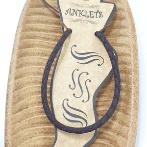 Könnte beinhalten: Ein braunes, geflochtenes Leder-Fußkettchen auf einem braunen, blattartigen Hintergrund mit einem Etikett, auf dem "ANKLETS" steht.
