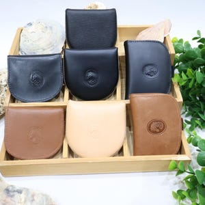 Può includere: Una collezione di portamonete in pelle in vari colori, tra cui nero, marrone e beige, esposti in un vassoio di legno. Ogni portamonete presenta un emblema circolare. I portamonete sono disposti con elementi decorativi.