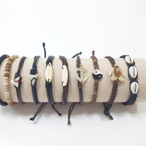 Peut inclure: Un ensemble de huit bracelets avec différents charmes, dont une coquille, une tortue, une ancre, un symbole yin yang et une dent de requin. Les bracelets sont en cuir marron, cordon noir et perles en bois.