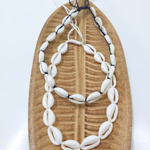 Könnte beinhalten: Zwei Halsketten aus weißen Muscheln, die auf einer schwarzen Schnur aufgefädelt sind. Die Halsketten werden auf einer braunen, blattartigen Oberfläche präsentiert.