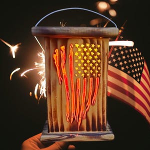 May include: A wooden lantern with an American flag design, featuring red stripes and star cutouts. The lantern has a handle and is illuminated, with sparks and an American flag in the background. A patriotic decoration.