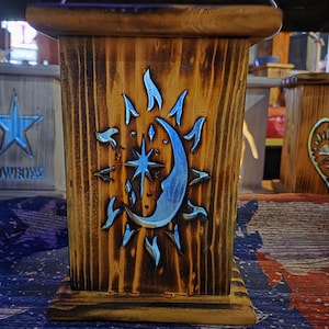 May include: A wooden box with a celestial design. The box is stained a warm brown and features a carved sun, moon, and star motif in silver. The design is centered on the front of the box, with a rustic, handcrafted appearance.