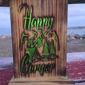May include: Wooden box with the words "Happy Camper" in green, along with a camping scene of a tent, trees, and mountains. The box has a natural wood grain finish and a rustic appearance.