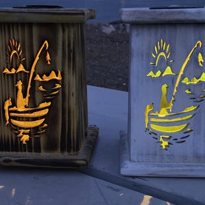 May include: Two wooden lanterns, one brown and one white, each featuring a cut-out design of a fisherman in a boat with a fishing rod, sun, and water. The cut-outs glow with a warm light, creating a cozy ambiance.