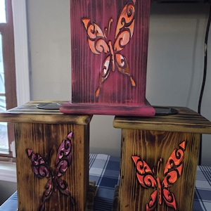 May include: Wooden candle holders with butterfly cutouts. The candle holders are stained in shades of brown and burgundy. The butterfly cutouts are painted in shades of orange and purple. The candle holders are square and have a flat top.