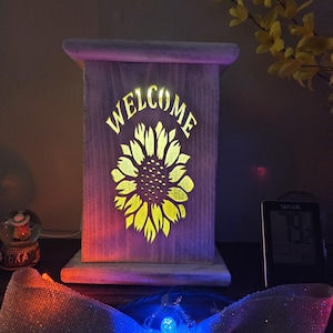 May include: Wooden welcome sign with a sunflower design. The sign is illuminated with a warm glow, featuring the word "WELCOME" at the top and a sunflower in the center. The sign is made of light-colored wood.