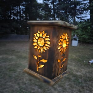 May include: A square wooden lantern with a dark, scorched finish featuring cut-out sunflower designs on its sides. The lantern emits a warm, glowing light from within and is suspended by a thin metal chain, showcasing visible wood grain.