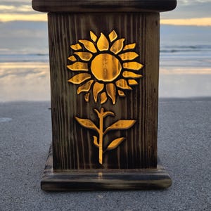 May include: A rectangular wooden lantern with a dark, scorched finish featuring a cutout of a sunflower on the front panel. The interior is illuminated, casting a warm golden glow through the sunflower shape, highlighting the wood grain texture.