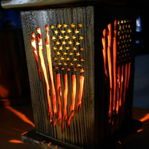May include: A square wooden lantern with a dark, weathered finish featuring a cutout design of the American flag. The stars and stripes are illuminated from within, casting a warm orange glow through the wood, with a metal handle on top.