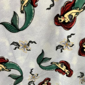 May include: A white fabric with a repeating pattern of Ariel, the Little Mermaid, in her mermaid form. She is depicted with red hair, a green tail, and a seashell bra. The background is a light blue with white flowers.