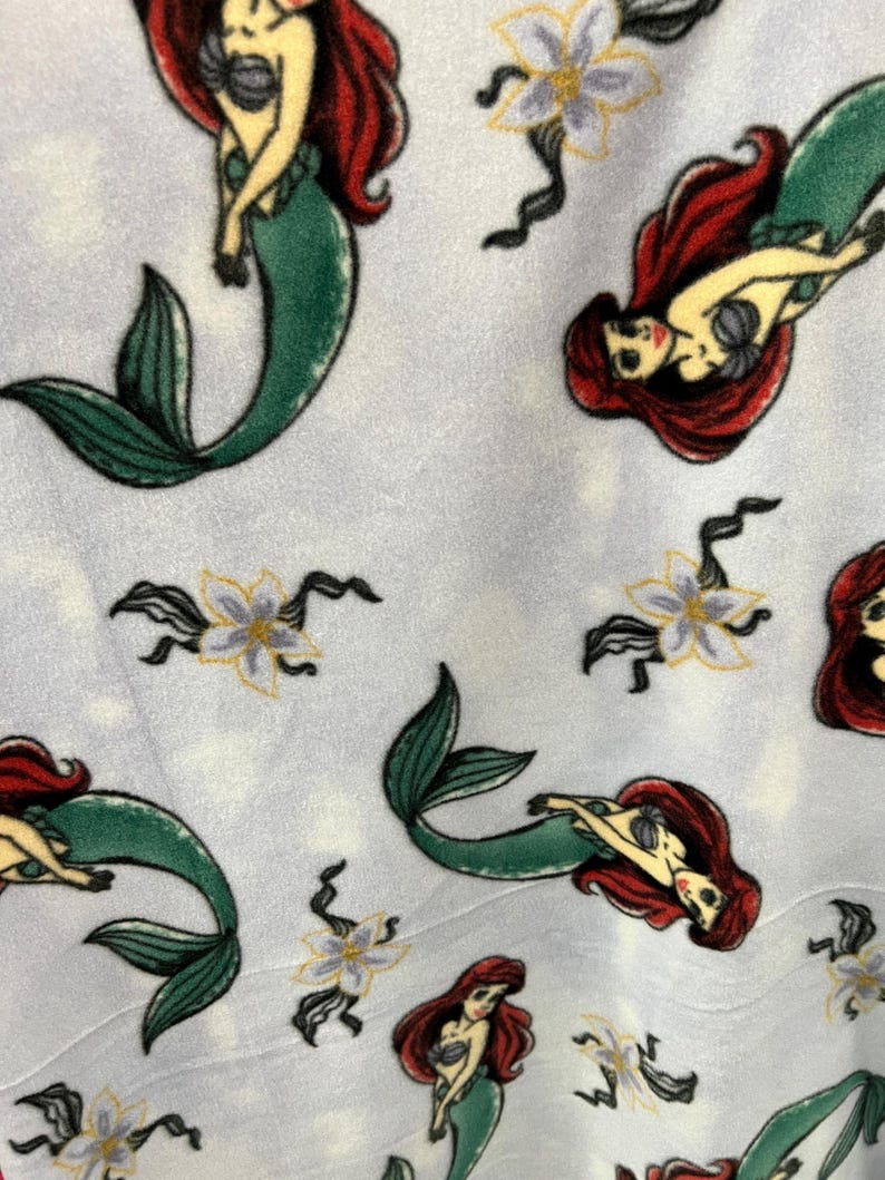 May include: A white fabric with a repeating pattern of Ariel, the Little Mermaid, in her mermaid form. She is depicted with red hair, a green tail, and a seashell bra. The background is a light blue with white flowers.