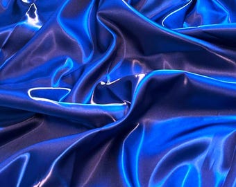 May include: A close-up shot of a vibrant, flowing fabric in shades of blue. The material has a glossy, satin-like sheen, with highlights and shadows creating depth. The fabric is draped and folded, adding texture and visual interest.
