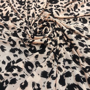 May include: Close-up of a fabric with a leopard print pattern. The fabric is a light beige color with black spots of varying sizes and shapes. The fabric is draped and folded, creating shadows and texture.