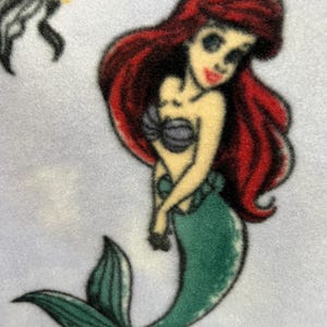 May include: A cartoon illustration of Ariel, the Little Mermaid, with long red hair, a green tail, and a purple seashell bra. The illustration is on a light blue background.