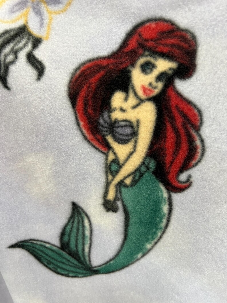 May include: A cartoon illustration of Ariel, the Little Mermaid, with long red hair, a green tail, and a purple seashell bra. The illustration is on a light blue background.