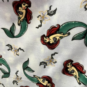 May include: A white fabric with a repeating pattern of Ariel, the Little Mermaid, as a mermaid with red hair and a green tail. The background is a light blue with white flowers.