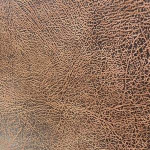 May include: Close-up of a textured surface resembling distressed leather. The colour is a blend of brown and black, with a network of fine, lighter brown lines creating a detailed pattern across the surface.