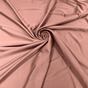 May include: A close-up shot of a swirl of dusty rose fabric. The fabric is smooth and appears to be a lightweight material, with soft folds and shadows creating depth. The color is a muted pink, reminiscent of blush.
