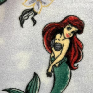 May include: A green mermaid with red hair and a seashell bra, on a light blue background with a white flower.