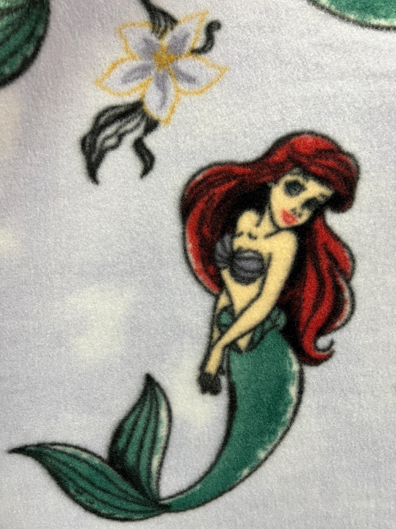 May include: A green mermaid with red hair and a seashell bra, on a light blue background with a white flower.