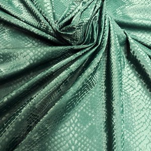 Puede incluir: Tela verde esmeralda con un patrón de piel de serpiente. La tela está doblada y drapeada, creando sombras y reflejos que enfatizan la textura y el brillo. El material tiene un acabado sutil y brillante.
