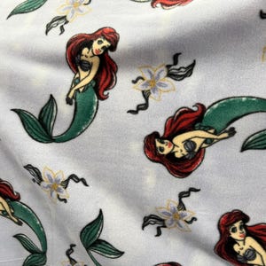 May include: A white fabric with a repeating pattern of a mermaid with red hair and a green tail. The mermaid is holding a seashell in her hand. There are also white flowers with yellow centers in the pattern.